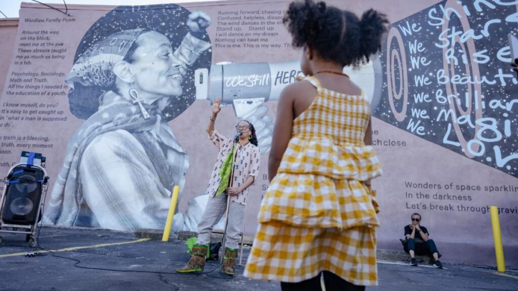 Young child standing in front of a mural and poet speaking into a microphone.